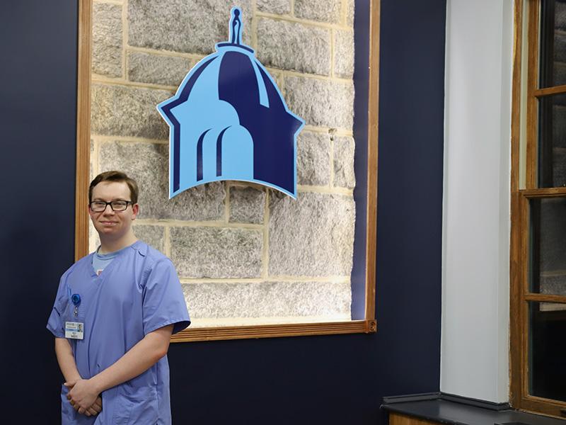 Student in scrubs standing in from of Immaculata logo